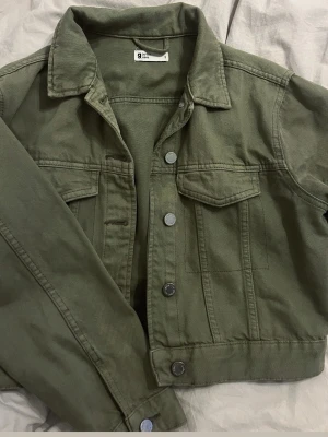 Olivgrön jeansjacka från Gina Tricot - Snygg olivgrön jeansjacka från Gina Tricot med klassisk krage, bröstfickor med lock och silverfärgade knappar. Jackan har en rak passform och är tillverkad i kraftigt jeanstyg. Perfekt för en cool och avslappnad stil.