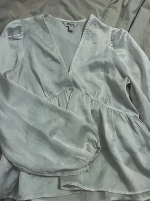 Vit v-ringad blus från Aware - Stilren vit blus från Aware med v-ringning och lätt glansigt tyg. Blusen har långa ärmar med diskret puff och resår vid ärmslut samt en lös, flowy passform. Perfekt för en trendig och avslappnad look.