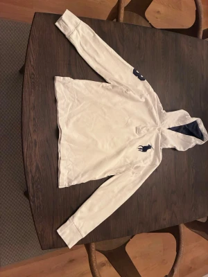 Vit långärmad skjorta från Ralph Lauren - Vit långärmad skjorta från Ralph Lauren med klassisk krage och mörkblå broderad logga på bröstet. Skjortan har knappar vid halsen och är tillverkad i ett mjukt bomullstyg. Perfekt för en stilren look. 10/12 år 152 cm