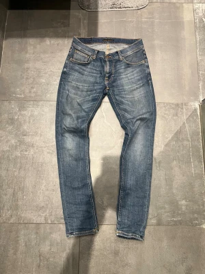 Blå jeans från Nudie Jeans - Säljer ett par klassiska blå jeans från Nudie Jeans med fem fickor och snygga slitningar. Jeansen har en rak passform och är tillverkade i denim med kontrastsömmar och Nudie-logga på bakfickan. Perfekta för en avslappnad och trendig look.