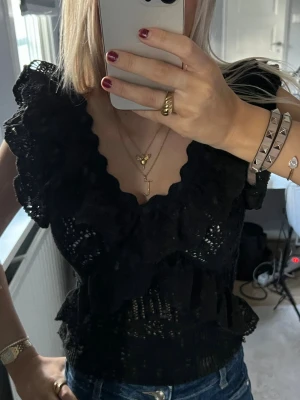 Svart volangblus med spetsdetaljer - Supersnygg svart blus med volanger och spetsdetaljer. Djup v-ringning och korta ärmar ger en trendig look. Perfekt att styla med jeans eller kjol för en cool vibe.