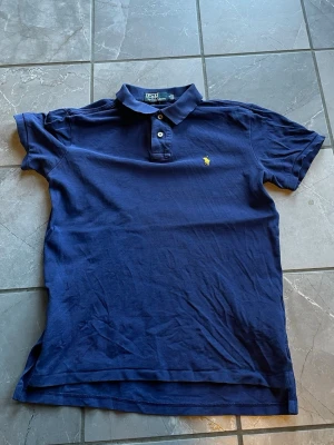 Marinblå pikétröja från Polo Ralph Lauren - Klassisk marinblå pikétröja från Polo Ralph Lauren med gul broderad logga på bröstet. Tröjan har krage, knappar framtill och korta ärmar. Perfekt för en stilren och sportig look. Vid snabb affär är jag villig att sänka priset något 
