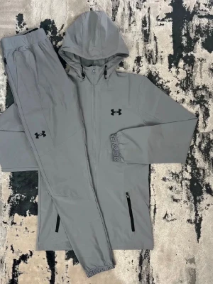 Grått loungewear-set från Under Armour - Snyggt grått loungewear-set från Under Armour med matchande hoodie och byxor. Hoodien har dragkedja, justerbar huva och svarta loggor på bröstet och byxan. Byxorna har resår i midjan och dragkedjeförsedda fickor. Perfekt för chill dagar hemma.