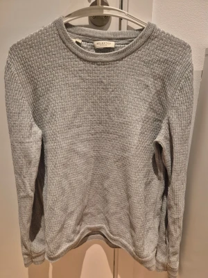 Grå stickad tröja Selected Homme S - Säljer en stilren grå stickad tröja från Selected Homme. Tröjan har rund halsringning, lång ärm och ett diskret vävt mönster i materialet. Perfekt för lager på lager och enkel att matcha med olika outfits. 100% bomull.