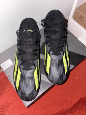 Adidas fotbollsskor svart/gul - Snygga fotbollsskor från Adidas med svart bas och skarpa gula ränder längs sidorna. Skorna har en modern, strömlinjeformad design med snörning och ovandel i syntetmaterial. Perfekta för dig som vill sticka ut på planen med både stil och funktion.