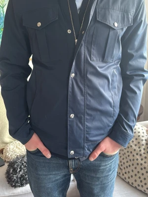 Mörkblå fieldjacket från J.Lindeberg - Stilren mörkblå fieldjacket från J.Lindeberg med silverfärgade knappar och dragkedja. Jackan har två bröstfickor med lock och knapp samt en diskret logotyp på ärmen. Perfekt för dig som gillar en klassisk look och perfekt nu inför våren