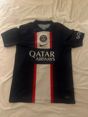 PSG matchtröja Nike blå/röd - Säljer en officiell Paris Saint-Germain matchtröja från Nike. Tröjan är mörkblå med röda och vita detaljer, PSG-logga på bröstet och Qatar Airways som sponsor. Korta ärmar och sportig passform, perfekt för fotboll eller träning. Köpt i PSG shoppen.