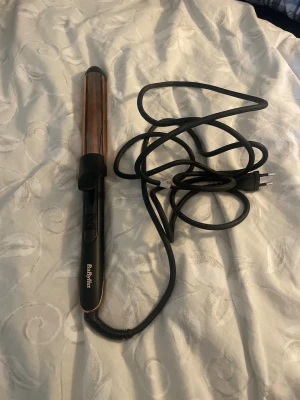 Svart locktång från BaByliss - Säljer en svart locktång från BaByliss med gulddetaljer och lång sladd. Tången har en cylinder i kopparfärg och enkel knappkontroll på handtaget. Perfekt för att fixa snygga lockar snabbt och smidigt.