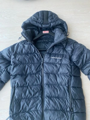 Svart dunjacka från Peak Performance - Snygg svart dunjacka från Peak Performance med huva och dragkedja framtill. Jackan har två sidofickor med dragkedja och diskret logga på bröstet. Perfekt för kalla vinterdagar och riktigt skön att bära.