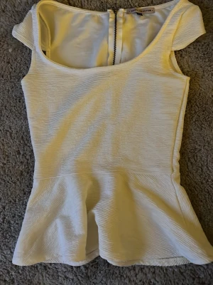 Vit topp med urringning och volang - Säljer en vit topp från berska COLLECTION med korta ärmar och rund halsringning.  har en markerad midja och dragkedja i ryggen för enkel påklädning. Materialet är strukturerat och ger en snygg formad siluett. Den är vit/beige