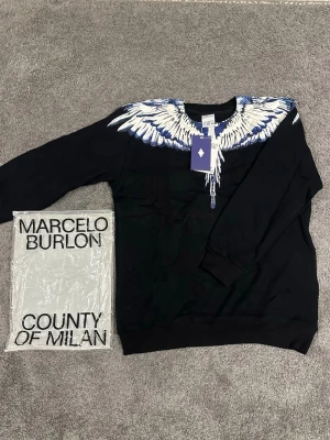 Marcelo Burlon tröja  - Svart tröja från Marcelo Burlon County of Milan med ikoniskt vingtryck i vitt och blått över axlar och bröst. Rund halsringning och långa ärmar. Tillverkad i mjuk bomull för en skön känsla. Perfekt statement-piece för dig som vill sticka ut.