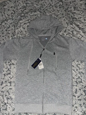  Polo Ralph Lauren hoodie - Snygg gråmelerad hoodie från Polo Ralph Lauren med dragkedja framtill och klassisk svart logga broderad på bröstet. Huva med dragsko och två fickor framtill. Perfekt för en chill och stilren look.