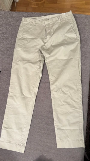 Beige chinos från Whyred - Snygga beige chinos från Whyred i klassisk rak modell. Tillverkade i bomull med sköna sidfickor och hällor för bälte. Perfekta för dig som gillar stilrena och enkla byxor med modern passform.