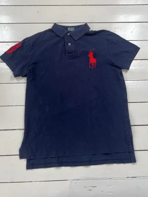 Mörkblå Ralph Lauren piké med röd logga - Klassisk mörkblå pikétröja från Ralph Lauren med stor röd broderad logga på bröstet och röd detalj på ärmen. Tröjan har korta ärmar, krage och knappar vid halsen. Perfekt för en stilren och sportig look.