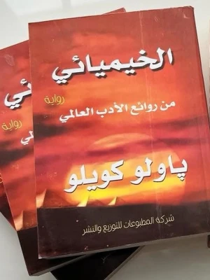 Alkemisten av Paulo Coelho på arabiska - En roman med titeln 'Alkemisten' av Paulo Coelho, översatt till arabiska. Boken har ett omslag i varma toner av orange och brunt med vit och gul text. Mjukpärm och klassisk skönlitteratur som inspirerar till äventyr och självupptäckt.