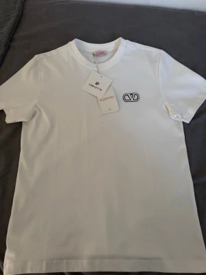 Vit t-shirt från Valentino - Vit t-shirt från Valentino med svart broderad logga på bröstet. Klassisk rund halsringning och korta ärmar. Tillverkad i mjuk bomull för en skön och stilren look. Perfekt för dig som gillar exklusiva och minimalistiska plagg.