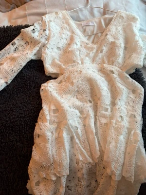 Vit spetsklänning med v-ringning - Superfin vit klänning med v-ringning och lång ärm. Klänningen har ett vackert spetsmönster och omlottkänsla upptill. Perfekt för sommarens alla tillfällen och har en romantisk vibe.