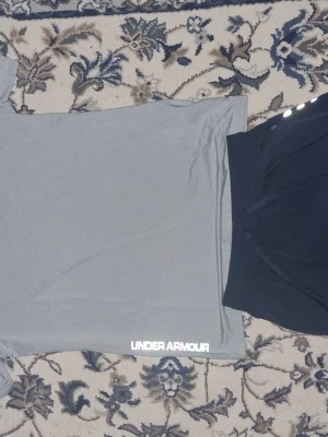 Grå träningströja från Under Armour - Snygg grå funktions-tshirt från Under Armour med korta ärmar och rund hals. Tröjan har vita logotyper och textdetaljer på bröstet och sidan. Tillverkad i ett lätt och snabbtorkande material som passar perfekt till träning.