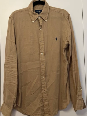Beige linneskjorta från Ralph Lauren - Klassisk beige skjorta från Ralph Lauren i linne med lång ärm och knappar framtill. Skjortan har button-down krage och den ikoniska mörkblå loggan broderad på bröstet. Perfekt för en avslappnad men ändå stilren look.