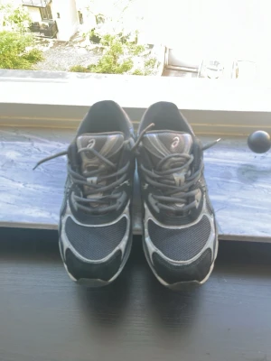 Svarta asics med meshade detaljer! - Säljer ett par svarta Asics sneakers med grå och vita detaljer. Skorna har en sportig design med meshpaneler för extra ventilation och klassiska snörningar. Perfekta för dig som gillar streetwear och bekväma skor till vardags. Snygg logga på sidan och tungspetsen.