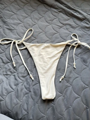 Vit bikiniunderdel med knyt och pärlor - Säljer en stilren vit bikiniunderdel med knyt i sidorna och dekorativa pärlor på snörena. Modellen är smal och högt skuren för en trendig look. Perfekt för stranden eller poolen i sommar!(tror dom är från Zalando)
