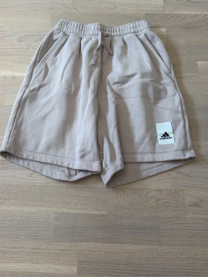 Beige shorts från Adidas med snörning - Snygga beige shorts från Adidas med elastisk midja och snörning. Två sidofickor och en liten Adidas-logga på vänster ben. Sköna och avslappnade, perfekta för sommardagar. Tillverkade i mjuk bomull för extra komfort.