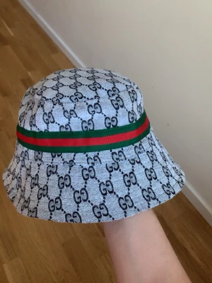 Gucci bucket hat med GG-mönster - Säljer en bucket hat från Gucci i ljusgrått tyg med klassiskt GG-monogram över hela hatten. Runt hatten går ett grönt och rött band. Insidan har ett coolt tigerprint. Perfekt för dig som vill sticka ut med en lyxig accessoar. Byten ingår, priset kan diskuteras!!