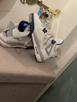 Nike Air Jordan 4 Retro Vit/Blå - Säljer ett par Nike Air Jordan 4 Retro sneakers i vitt läder med grå och marinblå detaljer. Skorna har meshpaneler, synlig Air-sula och klassiska Jordan-loggor på häl och plös. Perfekta för dig som gillar streetwear och ikoniska modeller.
