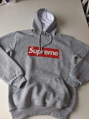 Grå Supreme hoodie med röd logga - Snygg grå hoodie från Supreme med den klassiska röda boxloggan på bröstet. Hoodien har huva med dragsko, magficka och ribbade muddar. Perfekt för dig som gillar streetwear och vill sticka ut med en ikonisk look. Den är också i mycket bra skick i storlek S. Hör av dig vid frågor