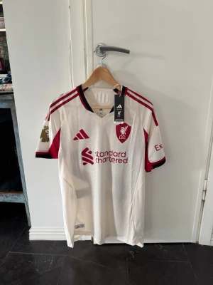 Liverpool bortatröja Adidas Ekiti 22 - Snygg Liverpool FC bortatröja från Adidas i vitt med vinröda detaljer och svarta inslag. Tröjan har tryck med Ekitike och nummer 22 på ryggen, samt Premier League-märke på ärmen. Materialet är väldigt lätt. 100% äkta, köpt på Liverpools hemsida och kvitto finns. (Aldrig använt)
