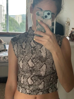 Ärmlös topp med ormmönster - Säljer en croppad, ärmlös topp med hög hals och snyggt ormmönster i svart och grått. Toppen har en tight passform och är gjord i ett mjukt, stretchigt material som känns bekvämt mot huden. Perfekt för dig som vill sticka ut lite extra.