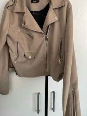 Beige skinnjacka från ONLY - Snygg beige skinnjacka från ONLY med klassisk bikerstil. Jackan har flera silverfärgade dragkedjor, axelklaffar och en liten ficka med knapp framtill. Kortare modell som ger en cool och trendig look.