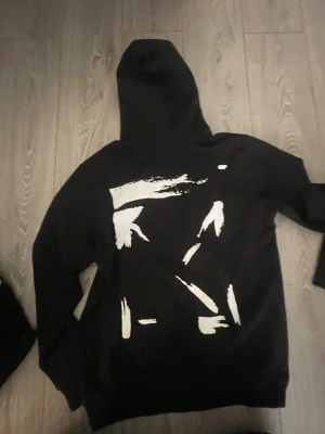 Svart hoodie från Off-White - Snygg svart hoodie från Off-White med vita penseldrag på ryggen och liten logga framtill. Klassisk huva och magficka. Perfekt för dig som gillar streetwear och vill sticka ut med en unik design.