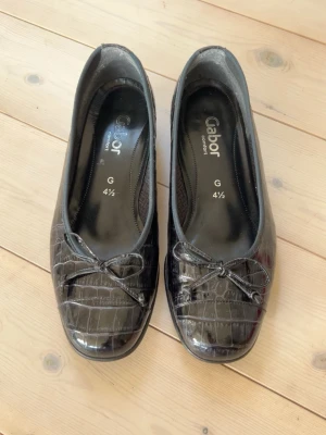 Svarta ballerinaskor från Gabor - Snygga svarta ballerinaskor från Gabor med lackad krokodilstruktur och liten rosett framtill. Skorna har rundad tå och låg klack, vilket ger en klassisk och stilren look. Perfekta för dig som gillar en elegant touch till outfiten.