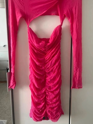 Rosa omlottklänning med cut out - Trendig rosa kortklänning med omlottdetalj och cut out över bysten. Klänningen har långa ärmar, rynkade detaljer framtill och figurnära passform. Perfekt för dig som vill sticka ut med en färgstark och modern look.