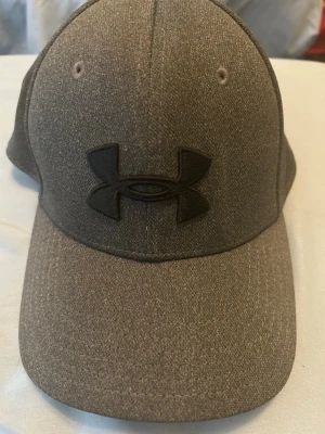 Mörkgrå keps från Under Armour - Snygg mörkgrå keps från Under Armour med böjd skärm och broderad svart logga framtill. Klassisk sexpanelsdesign med ventilationshål och justerbar passform. Tillverkad i ett mjukt tyg som ger en sportig och stilren look.