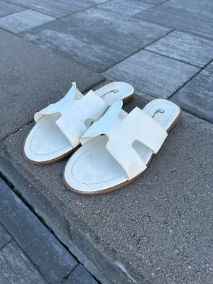 Vita slip-in sandaler från bambi  - Säljer ett par vita slip-in sandaler från Chiquelle med breda remmar i konstläder och öppen tå. Sandalerna har en klassisk design med en sula i beige och brunt samt en stilren look som passar till många outfits.
