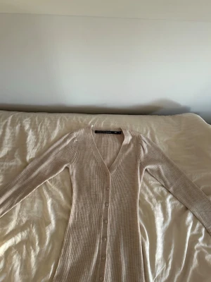 Beige ribbad långklänning med knappar aldrig använd - Säljer en beige ribbad långklänning med v-ringning och långa utsvängda ärmar mot slutet. Klänningen har knappar hela vägen framtill och är i ett mjukt, stretchigt material som sitter snyggt på kroppen. Perfekt för dig som gillar stilrena och bekväma plagg. 