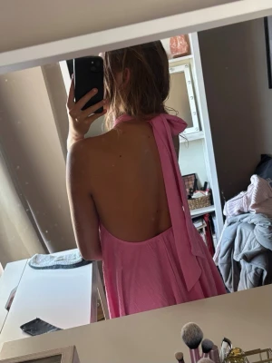 Rosa backless klänning som knyts i nacken -  Rosa klänning med  öppen rygg. Klänningen slutar vid lite längre ned än mitten på låren. Klänningen har halterneck med breda band som knyts i nacken och ett luftigt, svepande fall. Perfekt för sommarens alla tillfällen!