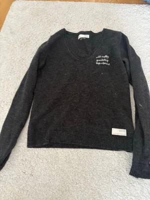 Mörkgrå stickad tröja från Zadig & Voltaire - Mörkgrå stickad tröja med V-ringning från odd molly . Tröjan har broderad text på bröstet  och dragkedjor vid ärmsluten. Perfekt till alla outfits.