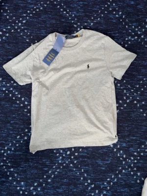 Grå t-shirt från Polo Ralph Lauren - Klassisk grå t-shirt från Polo Ralph Lauren med rund halsringning och korta ärmar. Tillverkad i mjuk bomull och har den ikoniska lilla broderade loggan på bröstet. Perfekt basplagg för en clean och stilren look. Storlek M men e liten i storlek passar som S