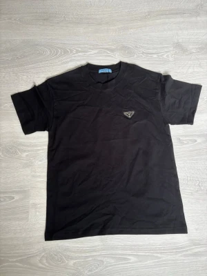 Svart t-shirt från Prada - Snygg svart t-shirt från Prada med klassisk passform och korta ärmar. T-shirten har en diskret logga på bröstet och är tillverkad i mjuk bomull som känns skön mot huden. Perfekt för dig som gillar stilrena och exklusiva plagg.