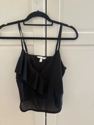 Svart volang topp från H&M - Snygg svart linnetopp från H&M med tunna axelband och volangdetalj framtill. Toppen har v-ringning och är tillverkad i ett lätt, tunt material som ger en luftig känsla. Perfekt för att skapa en trendig och stilren look.