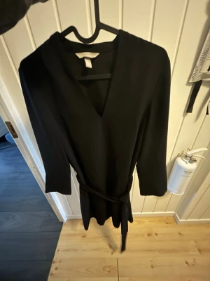 Svart omlottklänning med snöre - Stilren svart omlottklänning med v-ringning och knytband i midjan. Klänningen har långa ärmar och en klassisk, enkel design som passar till många tillfällen. Perfekt för dig som gillar minimalistisk stil. Använt några gånger 