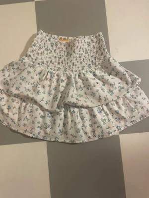 Vit blommig volang  - Supersöt kort kjol i vitt med småblommigt mönster i blått och rosa. Kjolen har smock i midjan och dubbla volanger för en extra fluffig look. I storlek 146/152 men eftersom de är stretchiga så passar de nog till en xxs😁Pris kan diskuteras🥰