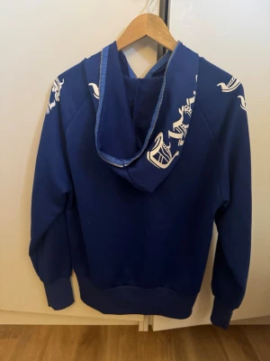 Blå zip hoodie med vita tryck - Säljer en blå hoodie med dragkedja från Kappa. Hoodien har vita Kappa-loggor längs ärmarna och på huvan, samt ribbade muddar vid ärmslut och nederkant. Perfekt för en sportig och avslappnad stil. Materialet känns mjukt och bekvämt.