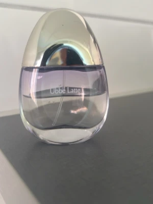 Zara Ubbe Latte parfym lila/silver - Säljer en parfym från Zara Ubbe Latte i en snygg oval flaska med silverfärgat lock och lila tonat glas. Flaskan har en modern och stilren design som känns lyxig och unik. Perfekt för dig som vill ha något som sticker ut på parfymhyllan.