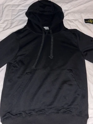 Svart hoodie från Stone Island - Snygg svart hoodie från Stone Island med klassisk huva och dragsnören. Hoodien har en stor magficka framtill och ribbade muddar vid ärmslut och nederkant. Tillverkad i mjukt bomullsmaterial som känns skönt mot huden. Perfekt för en avslappnad streetwear-look.