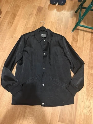 Svart overshirt från ASOS - Stilren svart overshirt från ASOS med klassisk krage och tryckknappar framtill. Jackan har två snedställda fickor och är tillverkad i ett slätt tyg som ger en clean look. Perfekt att slänga över en hoodie eller t-shirt för en enkel men snygg outfit.