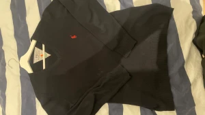 Mörkblå stickad tröja från Ralph Lauren - Mörkblå stickad tröja från Ralph Lauren med klassisk v-ringning och röd broderad logga på bröstet. Tröjan har långa ärmar och ribbstickade muddar vid ärmslut och nederkant. Perfekt för en stilren och avslappnad look.
